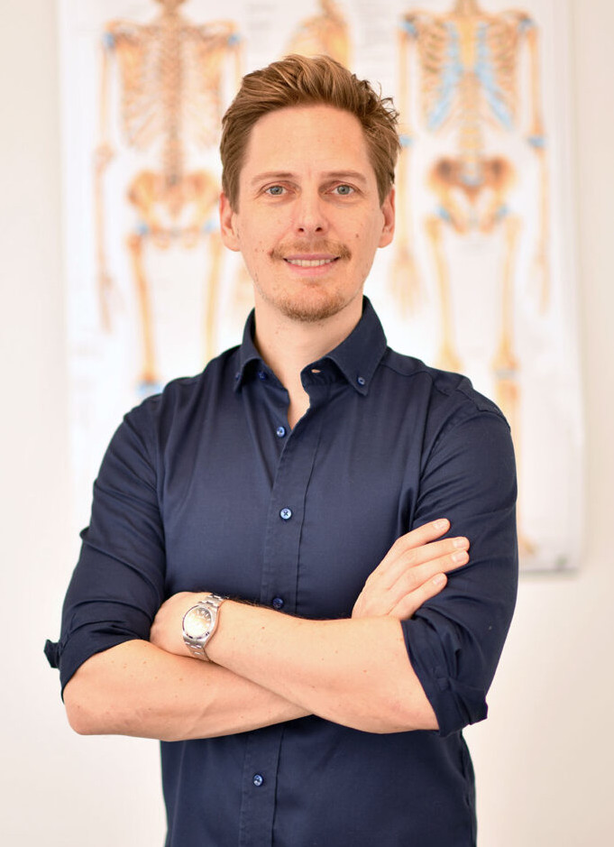 Sportarzt Dr. Stefan Treichler steht vor einer Wand mit anatomischem Bild eines Skelettes und lächelt mit locker verschränkten Armen.