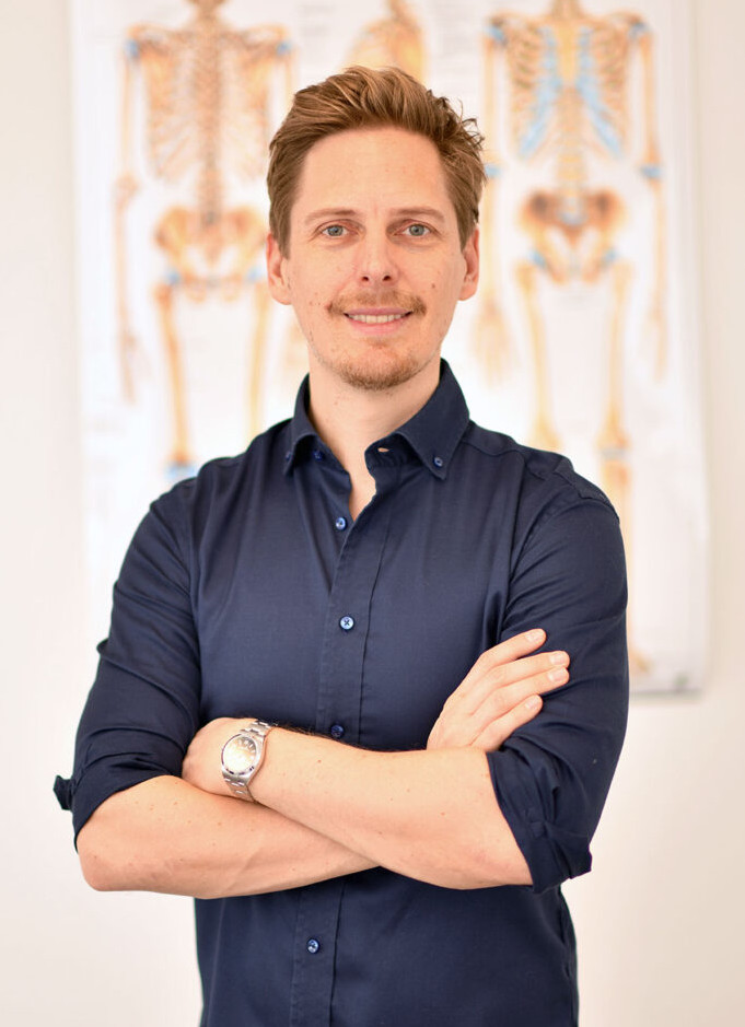 Manualmediziner Dr. Stefan Treichler steht vor einer Wand mit anatomischem Bild eines Skelettes und lächelt mit locker verschränkten Armen.