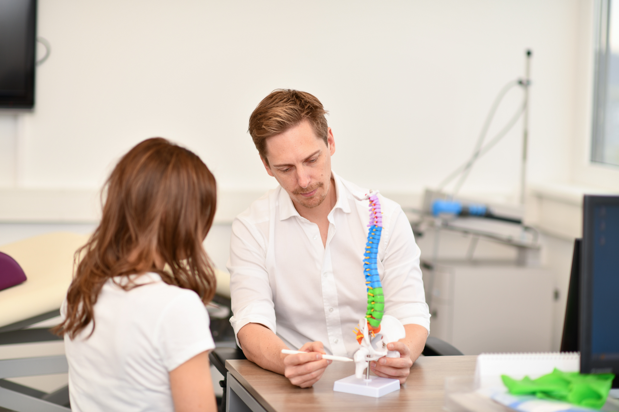 Dr. Treichler erklärt einer Patientin ihre Diagnose anhand eines Wirbelsäulenmodells und zeigt dabei mit einem Stift auf eine Stelle. 