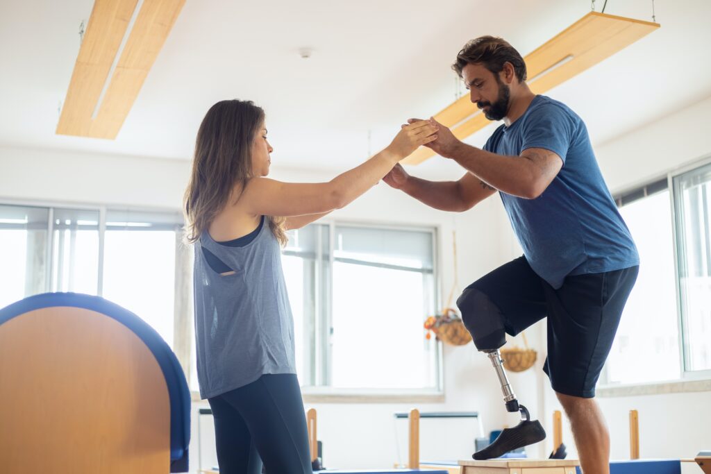 Mann mit Unterschenkelprothese wird beim Training des Einbeinstandes von einer Physiotherapeutin gehalten.