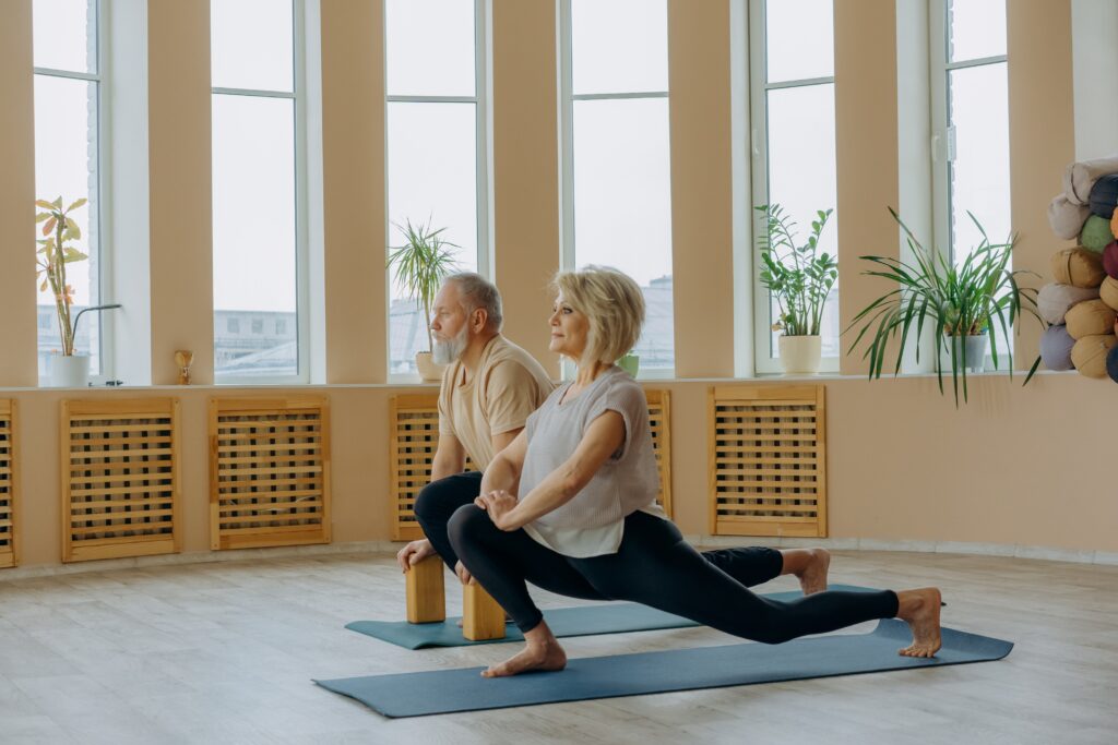 Eine ältere Dame und ein älterer Herr absolvieren einen Ausfallschritt. Training im Alter zum Erhalt der Leistungsfähigkeit.