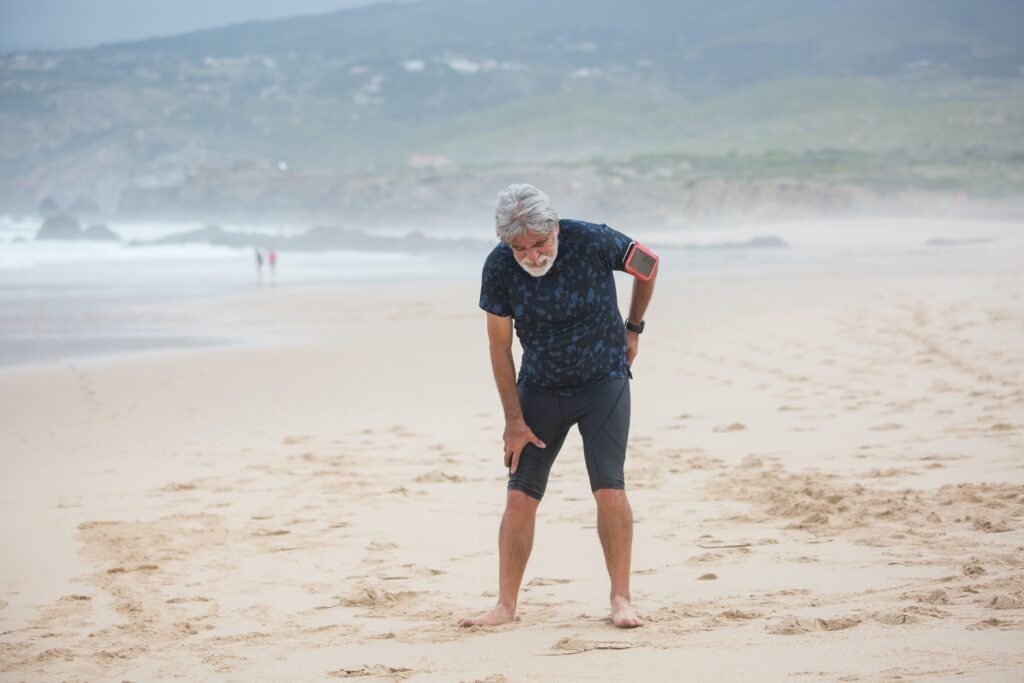 Ein älterer Mann steht am Strand und hält sich am Rücken als Zeichen für Schmerzen bei Osteoporose.